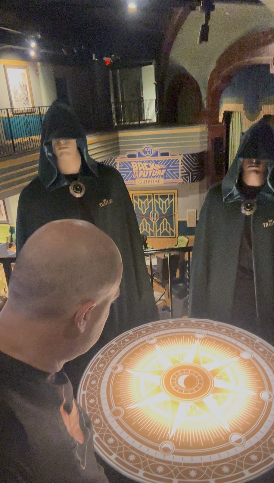 Ryan at a glowing round table between two hooded Traitors figures upstairs at Universal's Great Movie Escape